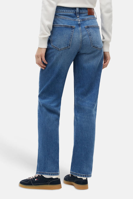 Midblauwe rechte jeans van TOMMY JEANS, achterzijde met klassieke zakken en bruin label, gedragen met witte trui en donkere sneakers.