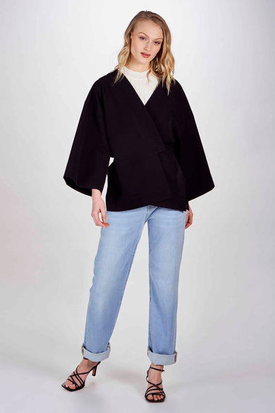 Blazer noir DEUX. par Eline De Munck, porté sur une chemise blanche et des jeans légers à talons.