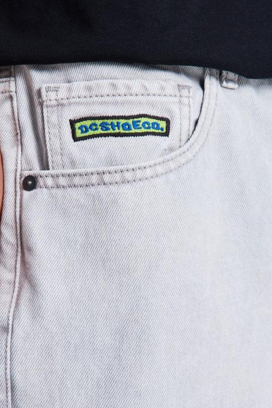 Lichtgrijze denim jeans met speciale pasvorm van DC SHOES, close-up van de voorzak met geborduurd logo en zwarte rivet.