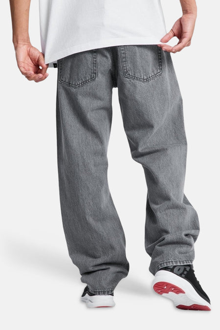 Midgrijze, wijdvallende spijkerbroek met speciale pasvorm van DC SHOES, met zichtbare achterzakken, gedragen met wit T-shirt en sneakers.