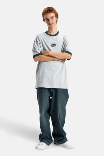 T-shirt gris à manches courtes de DC SHOES, avec des bordures vertes et des jeans décontractés et des chaussures blanches.