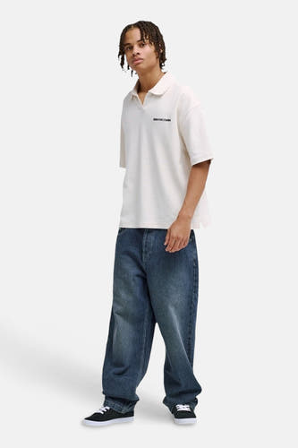 Polo blanche DC SHOES avec col ouvert et logo texte noir, portée par un homme avec un jean ample et des baskets noires.