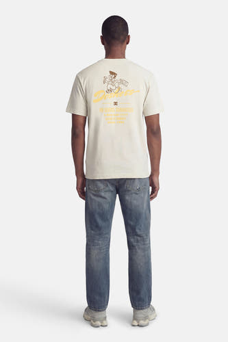 T-shirt beige de DC SHOES, avec un dessin animé et du texte au dos, porté avec un jean et des baskets grises.