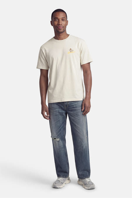 Beige T-shirt van DC SHOES met korte mouwen, losse pasvorm en kleine print op de borst, gedragen met blauwe jeans.
