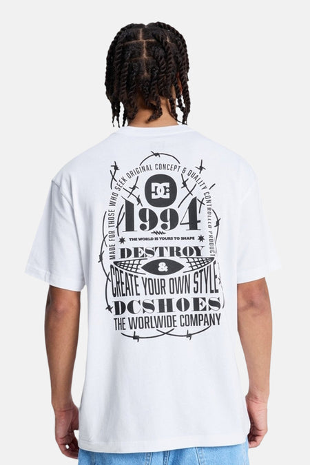 T-shirt blanc à manches courtes DC SHOES avec un grand imprimé noir au dos, incluant le logo DC, l'année 1994, des textes et des motifs de fil barbelé.
