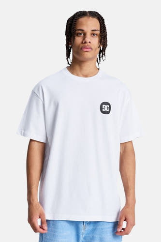 Wit T-shirt met korte mouwen van DC SHOES, met een zwart-wit logo op de linkerborst, gedragen door een man met gevlochten haar en een blauwe jeans.