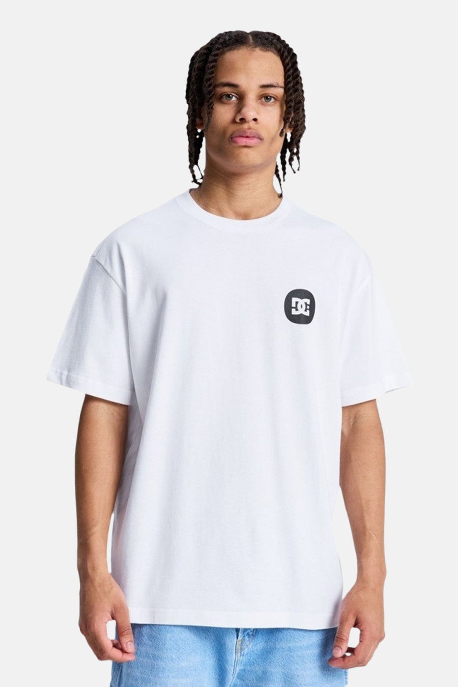 Wit T-shirt met korte mouwen van DC SHOES, met een zwart-wit logo op de linkerborst, gedragen door een man met gevlochten haar en een blauwe jeans.