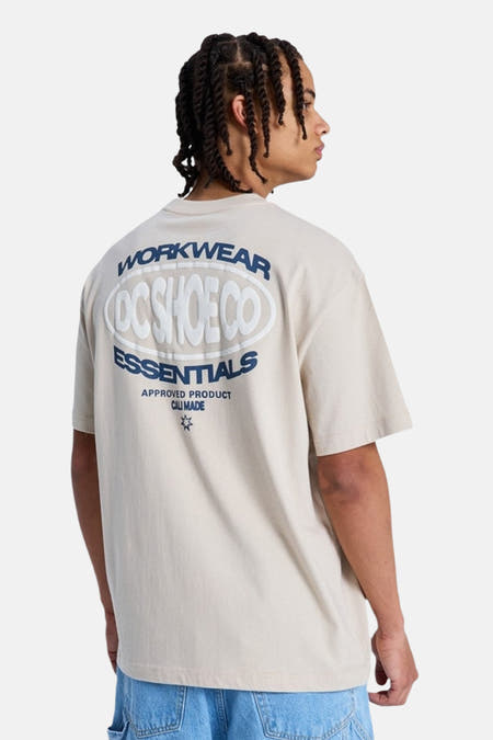 Beige T-shirt met korte mouwen van DC SHOES, met witte reliëf- en donkerblauwe tekstopdruk op de rug, gedragen door een persoon.