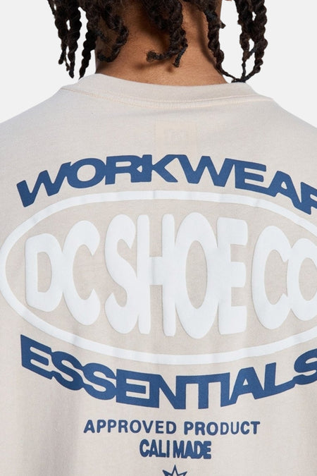 Beige T-shirt met korte mouwen van DC SHOES, achterkant met grote opdruk: witte, verhoogde 'DC SHOE CO' tekst in ovaal en blauwe 'WORKWEAR' en 'ESSENTIALS' teksten.