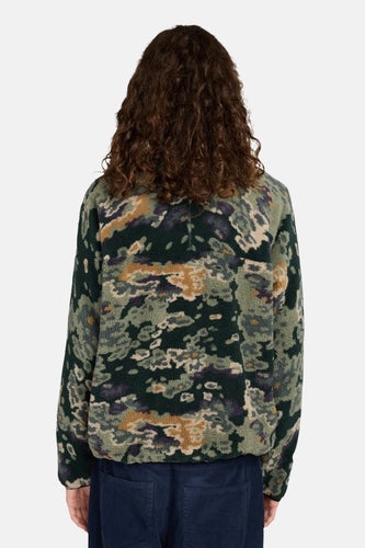 Persoon met krullend haar, camouflage fleece jas, marineblauwe broek.