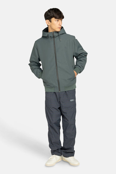 Korte groene jas van ELEMENT, met capuchon en zwarte ritssluiting, gecombineerd met geruite broek en witte schoenen.

