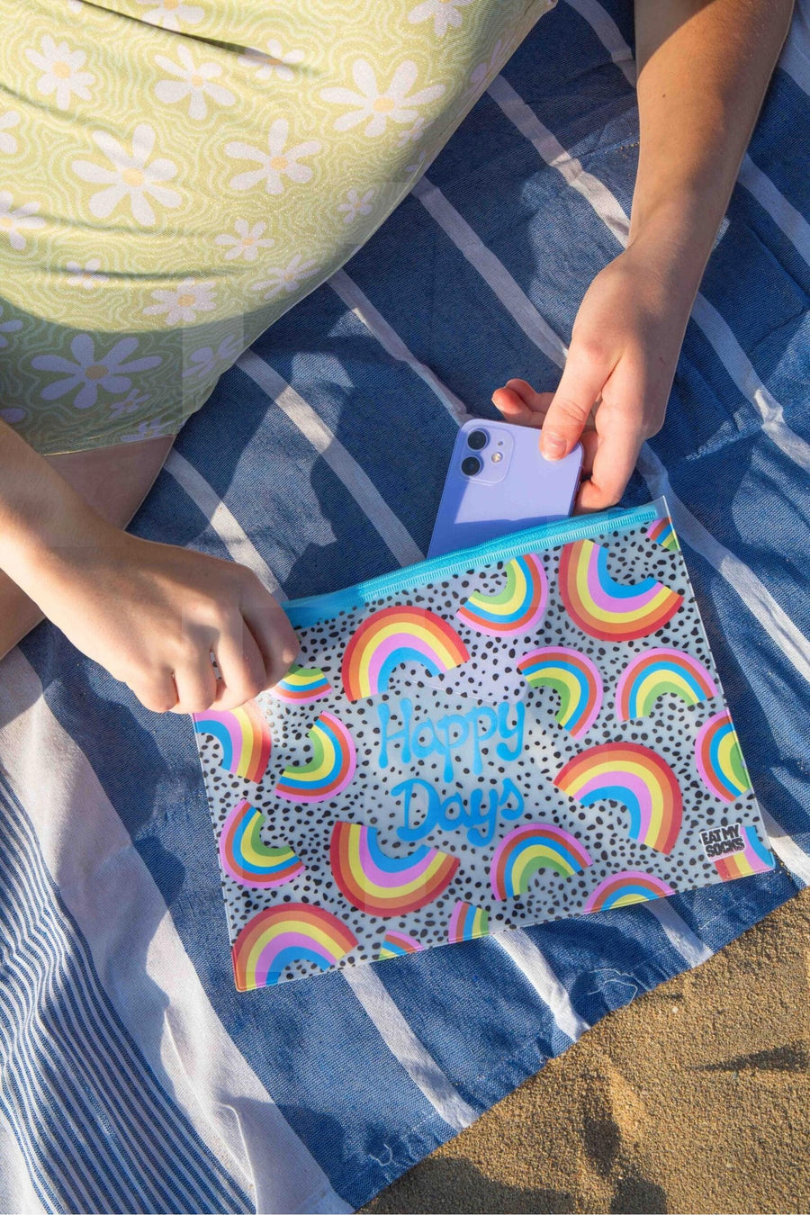 Multicolor toilettas met regenboogprint en "Happy Days" tekst van EAT MY SOCKS, op blauw gestreept strandlaken, close-up van hand die telefoon erin legt