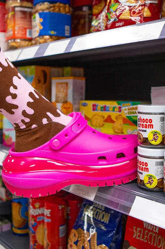Multicolor sokken met bruin-roze camouflagepatroon van EAT MY SOCKS, gedragen met felroze krokostijl schoenen, op supermarktplank met snoepjes.