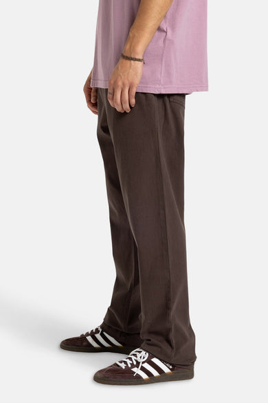 Pantalon brun de Quiksilver, avec des jambes droites, associé à un T-shirt mauve et à des baskets brunes avec des rayures blanches.