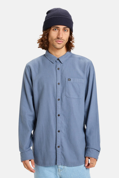 Chemise bleue à manches longues pour hommes de Quiksilver, avec une poche po poitrine et portée avec un chapeau bleu marine.