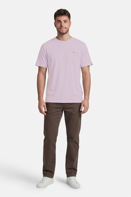 T-shirt violet de Quiksilver avec petit logo sur la poitrine, porté par un homme avec un pantalon marron et des baskets blanches.