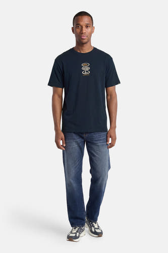 T-shirt à manches courtes bleu foncé Quiksilver, avec un imprimé graphique de couleur or et porté par un homme en jean et baskets.