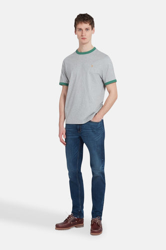 Man staat met een grijze T-shirt met groene boorden, blauwe jeans en bruine boots.