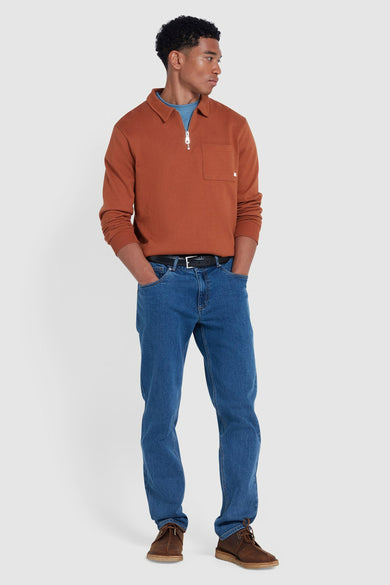 Man in bruine trui met rits, blauwe jeans en bruine schoenen staand.