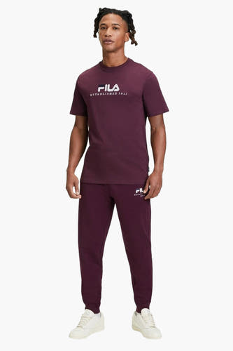 T-shirt rouge à manches courtes de FILA, avec une impression blanche sur l'avant, porté avec un pantalon de jogging.