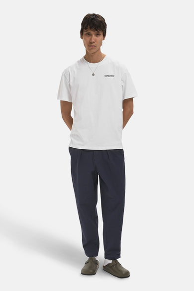 T-shirt blanc à manches courtes de POMPEII, porté avec un pantalon bleu marine et des chaussures vertes avec des chaussettes blanches.