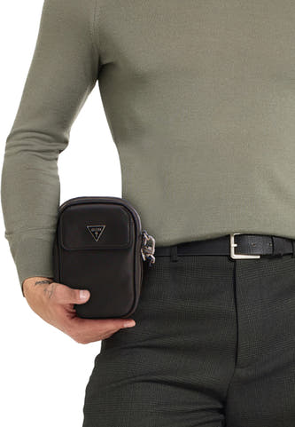 Zwarte schoudertas van Guess, met een zilverkleurig logo en een ritssluiting, gedragen met een olijfgroene trui en geruite broek.

