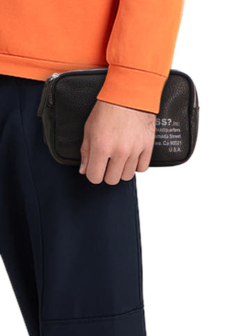 Zwarte schoudertas van Guess, met zachte textuur, gedragen met een oranje trui en donkerblauwe broek.