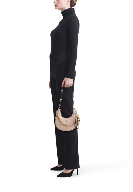 Beige schoudertas van Guess met gouden kettingdetails en zwarte merkbedel, gedragen door een vrouw in zwarte coltrui en pantalon.
