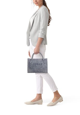 Blauwe handtas van Guess, met logo en korte handvatten, gedragen door een model met een grijze blazer en witte broek.
