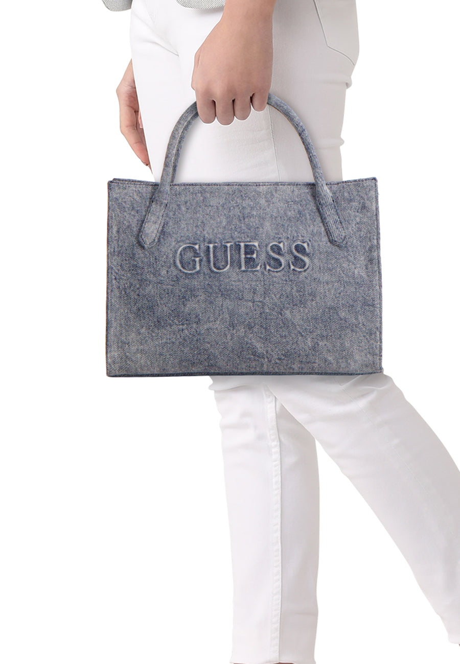 Blauwe handtas van Guess met de merknaam in reliëf en korte handvatten, gedragen bij een witte broek.
