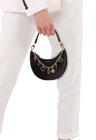 Zwarte schoudertas van Guess® met goudkleurige ketting en verschillende bedels, gedragen door een persoon in een wit pak.
