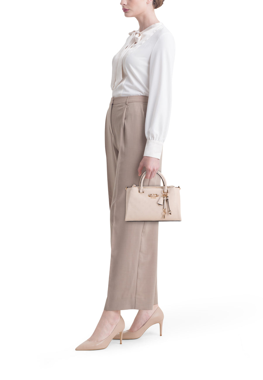 Sac à main beige Guess, avec un motif monogramme et des détails dorés, porté par un mannequin avec un chemisier blanc et un pantalon marron.