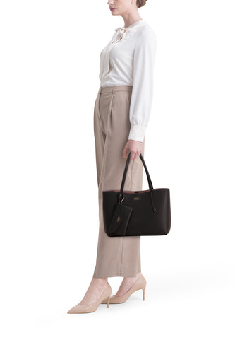 Vrouw met witte blouse, beige pantalon, bijpassende pumps en zwarte Guess handtas, staand tegen witte achtergrond.
