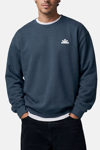 Pull bleu de BASELAB à col rond avec un petit logo blanc sur la poitrine, porté avec un t-shirt blanc et un pantalon noir.