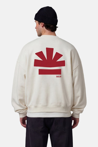 Pull blanc de BASELAB, avec imprimé rouge dans le dos, porté avec un bonnet noir et un pantalon noir ample.