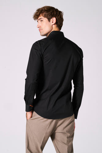 Chemise noire à manches longues de Le Fabuleux Marcel de Bruxelles, avec un pantalon marron et regardant vers la gauche.