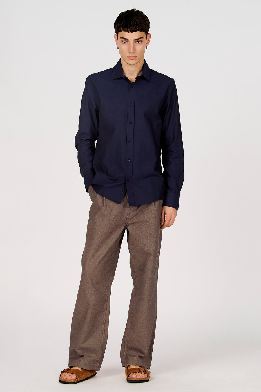 Chemise bleue à manches longues de Le Fabuleux Marcel de Bruxelles, portée avec un pantalon marron et des sandales.