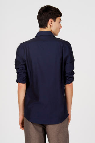 Chemise bleue à manches longues de Le Fabuleux Marcel de Bruxelles, vue de dos avec les manches retroussées et un pantalon marron.