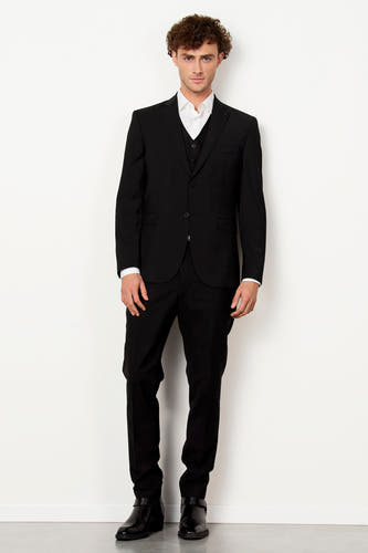 Blazer noir de Le Fabuleux Marcel de Bruxelles, avec des chaussures marrons et un collier, devant un fond blanc.