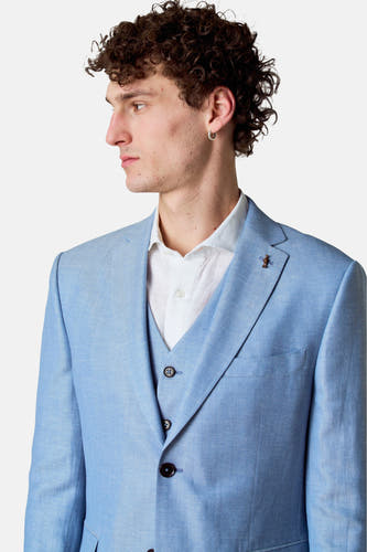 Blazer bleu de Le Fabuleux Marcel de Bruxelles, avec un motif subtil en maille de poisson, porté sur une chemise et un vest blanc.
