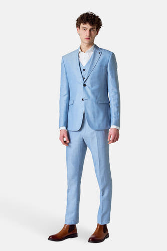 Gilet bleu de Le Fabuleux Marcel de Bruxelles, porté sous un manteau bleu clair avec une chemise blanche et des bottes brunes.