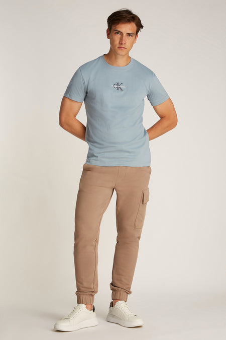 T-shirt bleu clair à manches courtes de Calvin Klein Jeans, porté avec un pantalon de jogging marron à poches et des baskets blanches.