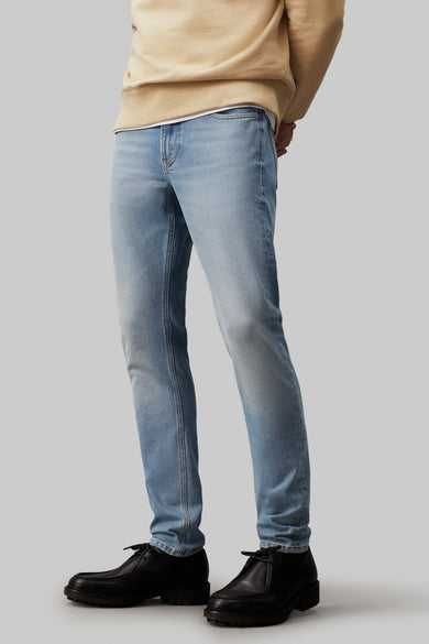 Slim tapered jeans van Calvin Klein, in lichtblauw denim, met zwarte schoenen en beige trui.
