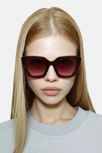Lunettes de soleil œil de chat rouge foncé de KOMONO, portées par une femme aux cheveux blonds et portant un t-shirt gris clair.