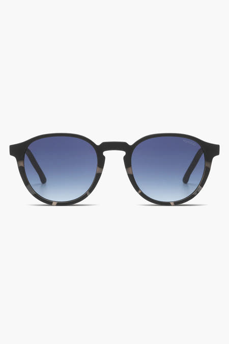 Lunettes de soleil noires de KOMONO, portées par une femme avec un foulard noir et un t-shirt bleu à imprimé batik.