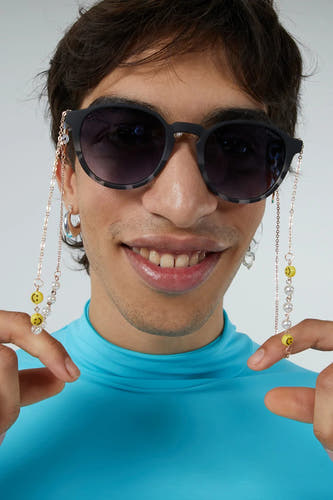 Zwarte zonnebril van KOMONO, met een ketting met parels en smiley-gezichtjes, gedragen met een blauwe coltrui.
