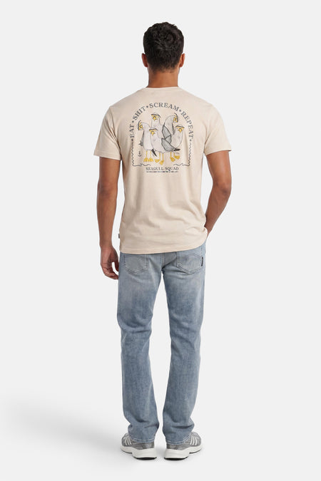 Ecru T-shirt van LAKOR met een 'Seagull Squad' print op de achterkant, gedragen door een model in jeans en sneakers.