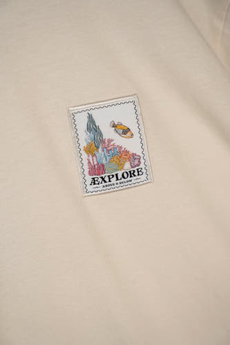 Ecru T-shirt met korte mouwen van LAKOR, met een vierkante patch met onderwatermotief en de tekst "Explore".
