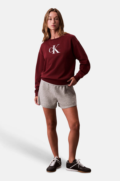 Vrouw draagt bordeauxrode Calvin Klein trui, grijze shorts, zwarte sneakers, staand.
