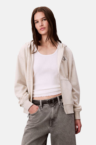Veste beige de Calvin Klein, portée sur un chemisier blanc et des jeans gris avec ceinture.